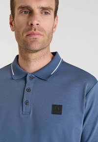 Polo shirt blu in cotone, con colletto a righe bianche, patta con tre bottoni e un'etichetta logo nera sul petto.