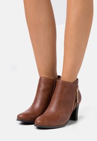 Bottes bottines en cuir synthétique marron avec un bout arrondi et une fermeture éclair latérale. Elles possèdent un talon épais de 3 pouces et une texture lisse.
