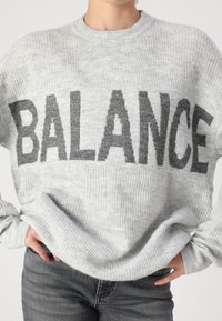 Szürke kötött pulóver, sötétebb szürke "BALANCE" grafikai mintával, bordás textúrával, kerek nyakkivágással és enyhén bő szabással.