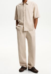 Camicetta beige in lino a maniche corte con chiusura a bottoni e pantaloni abbinati a vestibilità ampia. Indossata con scarpe marroni scure. Texture morbida, design minimalista.