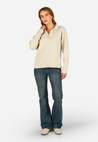 Krémszínű pulóver, vaffel struktúrával, gallérral és cipzárral. Kék, trapéz szabású farmerrel és krémszínű cipővel párosítva.