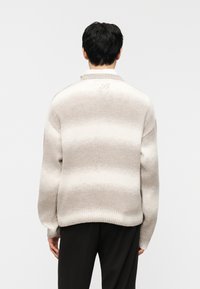 Homme aux cheveux noirs courts portant un pull beige à rayures blanches, debout de dos devant un fond clair uni.