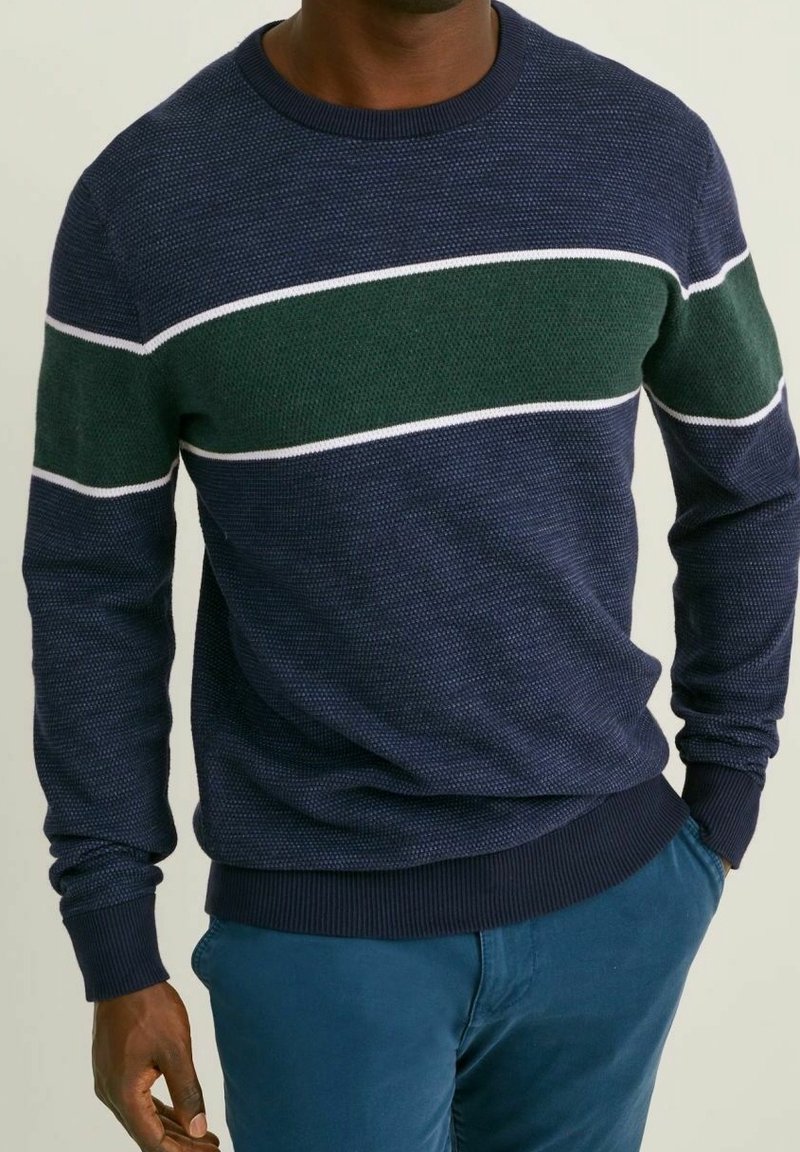 Stickad tröja i marinblått med en grön och vit horisontell rand över bröstet. Har rund halsringning och ribbade muddar.