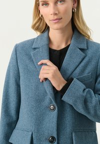 Hellblauer Wollmischblazer mit Reverskragen, zwei Knöpfen und einer Brusttasche. Der Stoff hat eine weiche Textur.