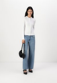 Cardigan blanc à manches longues avec des boutons, associé à un jean en denim bleu à jambes larges. Les accessoires comprennent un petit sac noir et des escarpins noirs pointus.