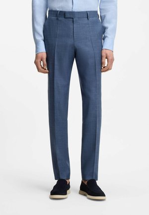 Hombre con pantalones de vestir azul ajustados, camisa azul clara metida por dentro y mocasines negros con suelas beige sobre fondo blanco.