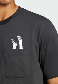 Μαύρη βαμβακερή μπλούζα με κοντά μανίκια, διαθέτει τσέπη στο στήθος με άσπρο κεντημένο σχέδιο ποδιών σε κάλτσες και παντόφλες. Στρογγυλή λαιμόκοψη.