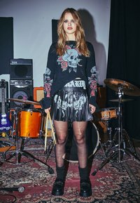 Junge Frau mit Drumsticks steht in einem Musikstudio mit Schlagzeug, Gitarren und Verstärkern, trägt einen Pullover mit Totenkopfprint und einem Rock mit gerissenen Strumpfhosen.