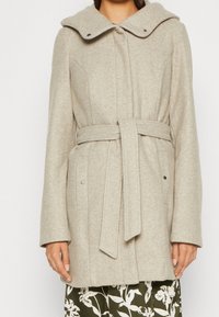 Cappotto in lana beige con ampio colletto, vita con laccio e tasche frontali. Presenta chiusure a bottone e un tessuto morbido e testurizzato.