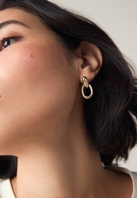 Gold hoop earrings with a linked design, featuring a shiny finish and smooth texture, worn on a model's ear against a neutral background.
