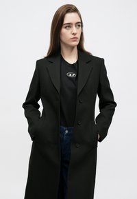 Juodas ilgas paltas iš struktūruotos medžiagos, su siūlėta atlapė, trim mygtukais ir šoninėmis kišenėmis, dėvimas ant juodos marškinėlių ir mėlynų džinsų.