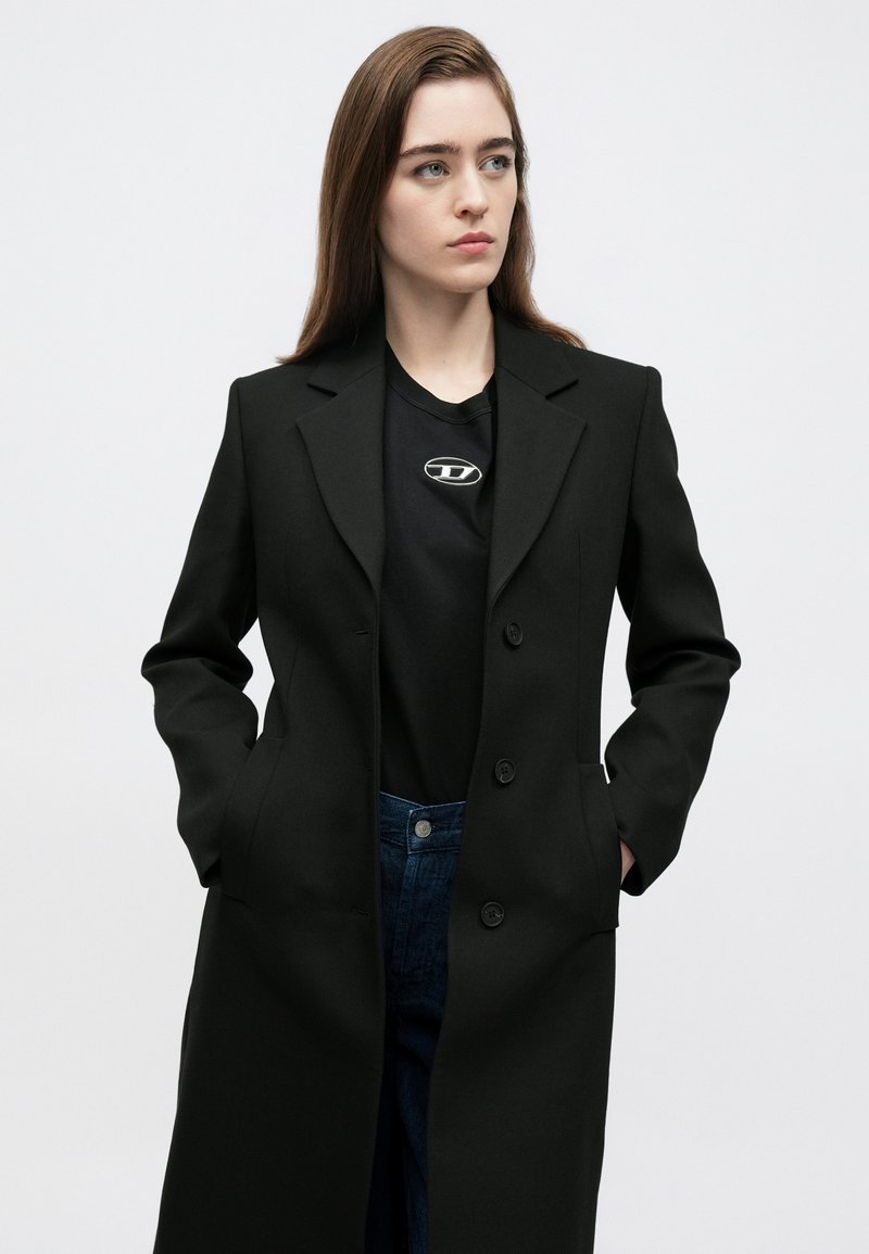 Sort lang frakke lavet af struktureret stof med en hakket revers, tre knapper og sidelommer, båret over en sort skjorte og blå jeans.