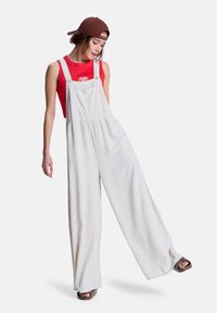 Salopette beige clair en tissu doux, avec jambes larges, bretelles ajustables et poche avant. Associée à un débardeur rouge et une casquette.