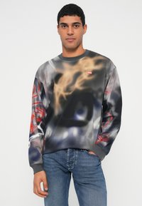 Νεαρός άνδρας που φοράει ένα φούτερ με πολύχρωμο αφηρημένο σχέδιο και λογότυπο Diesel, μαζί με μπλε τζιν, στέκεται μπροστά σε απλό φωτεινό φόντο.