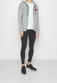 Uomo che indossa una felpa grigia con zip e logo rosso, una maglietta bianca, pantaloni neri aderenti e sneaker nere in piedi contro uno sfondo bianco.