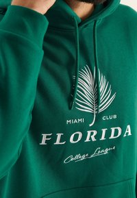Sudadera verde con un gráfico de hoja de palma y el texto "MIAMI CLUB" encima de "FLORIDA" y "College League" en bordado blanco. Textura suave.