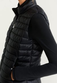 Gilet sans manches noir matelassé avec col montant et fermeture éclair. Il présente une texture lisse et légère ainsi qu'une coupe slim.