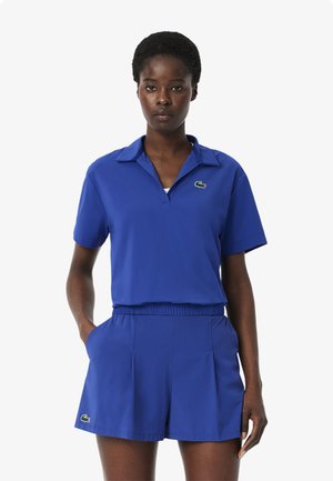 Blå polo-shirt med krave og korte ærmer, kombineret med matchende shorts med elastisk talje og sidelommer. Lacoste-logo som accent.
