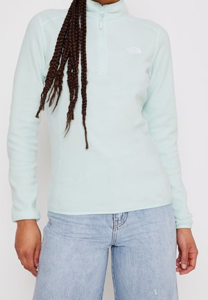 Femme portant une veste polaire à fermeture éclair bleu clair et un jean en denim clair, avec de longs cheveux tressés sur l'épaule.