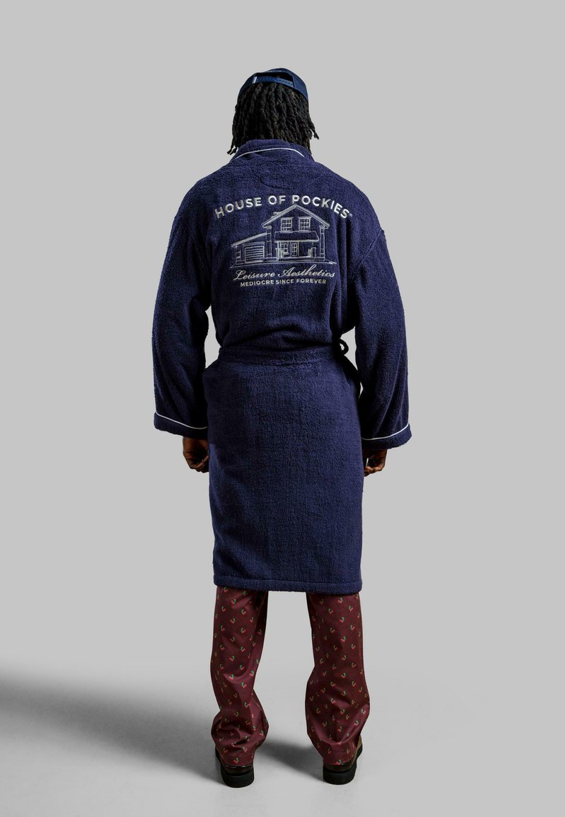 Marineblauwe badjas van badstof met witte bies, voorzien van een geborduurd huisgrafiek en tekst op de achterkant. Gepaird met gemêleerde bordeauxrode broek.