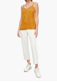 Top sans manches de couleur moutarde avec un tissu texturé et des bordures en dentelle. Associé à un pantalon court blanc et des baskets blanches.