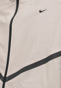 Κοντινή λήψη από το μπροστινό μέρος ενός ανοιχτό μπεζ μπουφάν με μαύρο φερμουάρ, μαύρη διαγώνια λωρίδα και μικρό μαύρο λογότυπο Nike Swoosh στο στήθος.