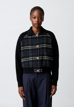 Donna che indossa una giacca a quadri nera e blu con bottoni, maniche nere e pantaloni blu navy a righe, in piedi davanti a uno sfondo semplice.