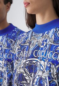 Blå grafisk sweatshirt med intrikate hvite blomster- og geometriske mønstre, ribbestrikkede mansjetter og rund hals med "CARLO COLUCCI" trykket foran.