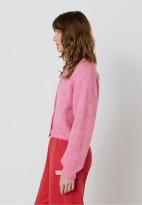 Ružová pletená cardigan s textúrovaným povrchom, s dlhými rukávmi a skráteným strihom. Nosí sa cez červené tepláky.