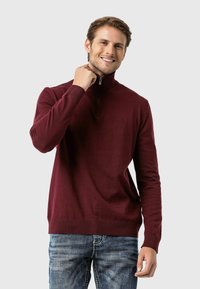 Burgunderfarbener Strickpullover mit hohem Kragen und Viertelreißverschluss, mit geripptem Saum und Bündchen, getragen über Jeans.