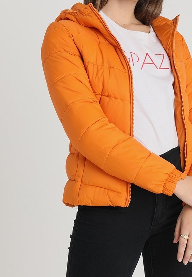 Orange pufferväst med luva, med ett quiltat mönster, dragkedja och sidofickor. Används över en vit t-shirt och svarta byxor.