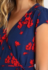 Kvinna iklädd en blå omslagsklänning med röda blommönster och korta ärmar, som visar överkroppen och en del av armen.