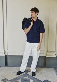 Homme en polo bleu marine et pantalon blanc portant un sac noir tressé sur l'épaule, debout sur un sol en carreaux à motif contre un mur beige.