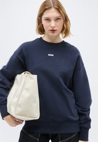 Γυναίκα με κοντά ξανθά μαλλιά, φορώντας μια μπλε μπλούζα BOSS, κρατώντας μια μεγάλη εκρού τσάντα ώμου μπροστά σε απλό φόντο.