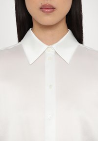 Witte blouse met lange mouwen en een satijnen afwerking. Voorzien van een klassieke puntige kraag en een knoopsluiting aan de voorkant, wat zorgt voor een elegante en gladde uitstraling.
