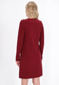 Abito lungo a maniche di colore bordeaux, in maglia strutturata, con bottoni decorativi sulle spalle e una linea dritta. Tessuto morbido con spacchi laterali.