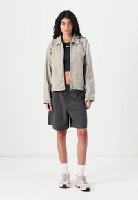 Light gray zip-up jacket, black crop top, patterned black shorts, and light sneakers. Model stands against a plain white background.