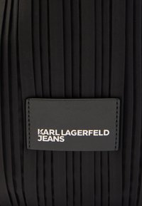 Czarny plisowany materiał z przyszytą prostokątną czarną metką, na której białymi wielkimi literami widnieje napis "KARL LAGERFELD JEANS".