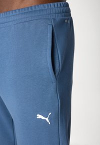 Blå bomullsoveraller med sidfickor, elastisk midja och en vit Puma-logotypaccents vid botten. Slät textur, avslappnad design.