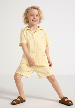 Gul kortärmad skjorta och shorts-set med grön palmträd-broderi, tillverkat av lättviktig tyg; parat med bruna sandaler.