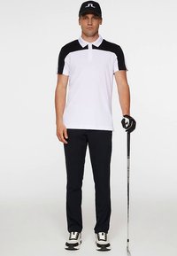 Vit och svart kortärmad pikétröja med tvåknappsknäppning, kombinerad med svarta byxor och svart golfkeps, håller i en golfklubba.