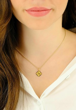 Collier en or avec un pendentif en forme de losange arborant un motif noir. La chaîne est fine et délicate, posée sur une chemise blanche.