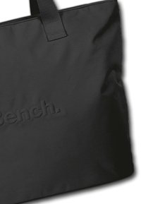 Sac fourre-tout noir en tissu lisse, avec de grandes poignées et un logo "Bench." embossé sur le devant. Design simple et minimaliste.