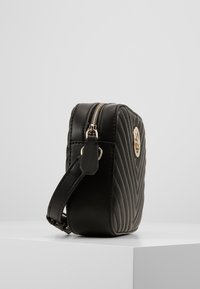 Sort quilted crossbody taske med guldfarvet lynlås og logo, vist på hvid overflade mod en lys grå baggrund.