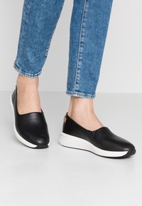 Chaussures noires à enfiler avec une tige en cuir lisse, semelle blanche et branding discret au talon. Portées avec un jean en denim bleu.