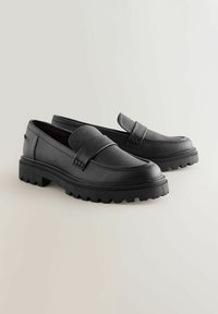 Mocasines de cuero negro con un acabado texturizado, que cuentan con una correa frontal y una suela de goma gruesa con un patrón de tracción robusto.