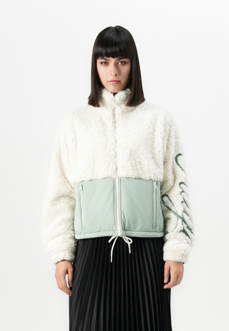 Femme avec des cheveux raides et foncés, portant une veste en polaire blanche avec des panneaux vert clair et une jupe plissée noire, se tenant devant un fond blanc.