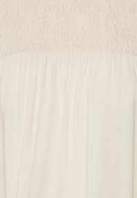 Krämlfärgad klänning med smockad överdel och flytande, veckad kjol. Mjuk, lättviktig tyg med en slät textur.