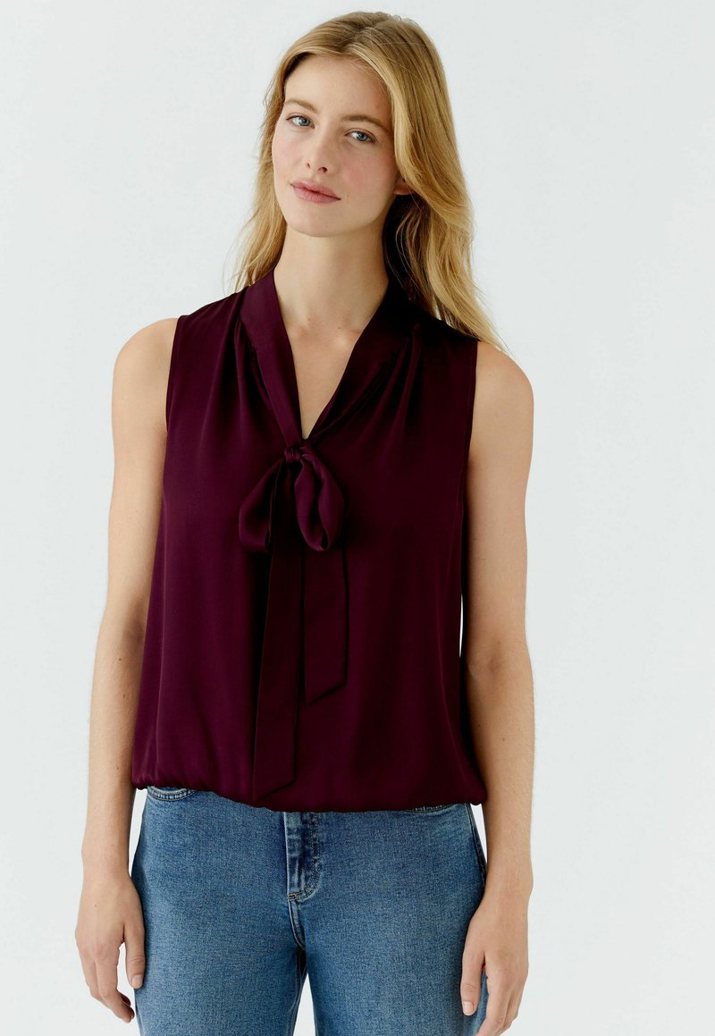 Bordeaux mouwloze blouse met een knoopdetail bij de hals, gemaakt van lichtgewicht stof, met een ontspannen pasvorm en subtiele glans. Jeans zichtbaar eronder.