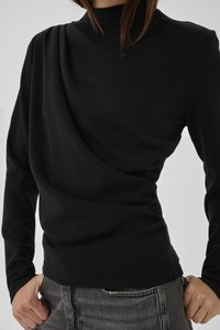 Žena koja nosi uski crni top s dugim rukavima s asimetričnim naborima i sive traperice, ruke blizu struka.